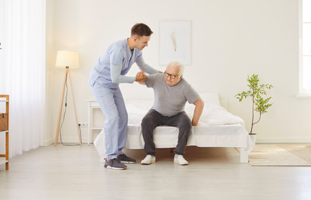 Portrait of a young friendly male nurse helping a senior patient to get off the bed at home. Physiotherapist or health care worker helping old man in rehab. Nursing service and support concept.の写真素材