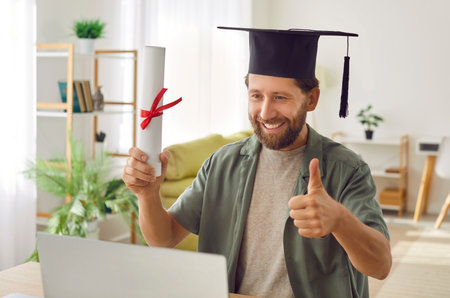 Young smiling graduate man having video call holding diploma in hands. Happy student sitting at the desk at home with laptop showing thumb up sign in a virtual ceremony. Online degree concept.の写真素材