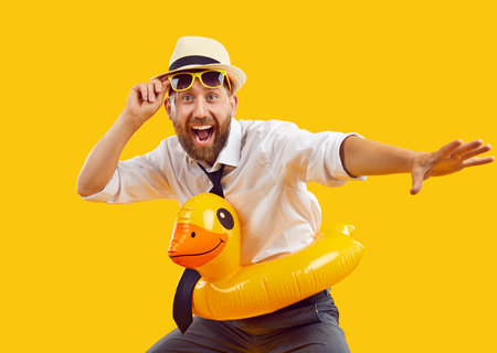 Happy excited office worker having fun on summer holiday. Studio portrait funny business man in white shirt, sunhat and rubber toy duck swim ring isolated on yellow orange background. Vacation conceptの写真素材