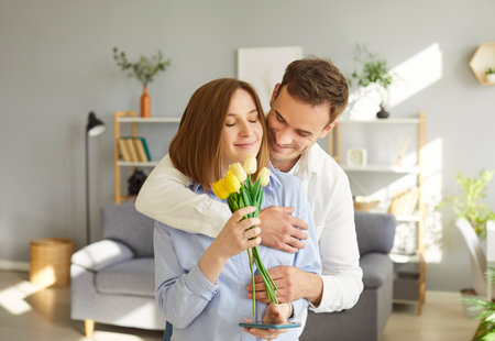 Loving husband gently hugging his wife, congratulating her with International Women Day, birthday or anniversary with bouquet of tulips. Happy couple celebrating romantic occasion together at home.の写真素材