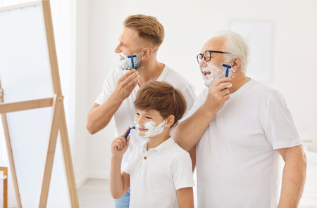 Grandfather, father and little son pretend to shave with cream on their faces, showing family bond. Three generations of men with shaving cream on faces hold razors and shave funny in front of mirror.の写真素材