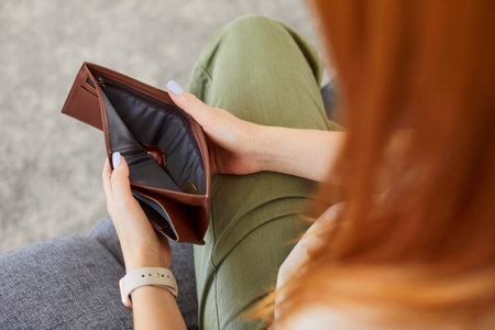 Young unemployed redhead woman holding opening empty wallet sitting on sofa in living room at home. Poor female person with no money. Bankrupt, financial crisis and poverty conceptの写真素材