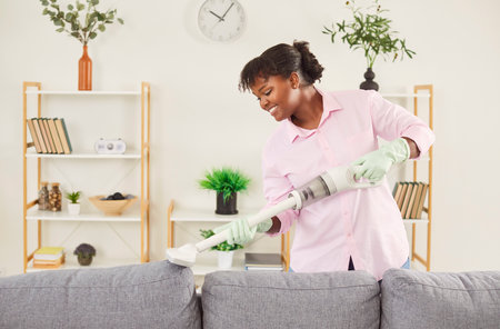 Happy smiling woman cleaning sofa with vacuum cleaner in the living room at home. Female janitor vacuuming furniture. Cleaning service, housekeeping, housework and household concept.の写真素材