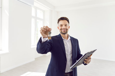 Portrait of confident smiling young man realtor agent standing in new white impartments with documents and house keys and looking cheerful at camera. Real estate market and moving day conceptの写真素材