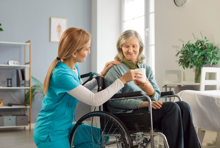Caring female doctor or nurse bringing tea to a senior woman patient sitting in a wheelchair at a...