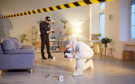 Photographing evidence, crime scene photography, investigator, policeman and professional criminological expert white coverall inspecting, examining behind barricade tape, yellow police line, safetyの写真素材
