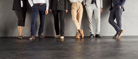 Legs of unrecognizable business people standing in a row in office on dark gray background. Cropped shot of a group of employees in formal clothes standing on a meeting. Teamwork concept. Banner.の写真素材