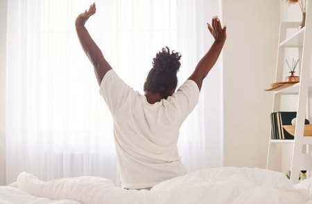 Back view of young African American woman in white t-shirt sitting in bed in front of window,...