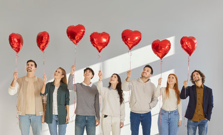 Portrait of a group of happy smiling people friends standing in a row and holding red balloon hearts in their hands. Young students standing on gray background. Love and friendship concept.の写真素材