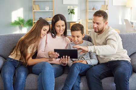 Portrait of a happy young cheerful family using tablet with children sitting on sofa at home. Mother and father enjoying weekend browsing internet with kids. Family leisure and technology concept.の写真素材