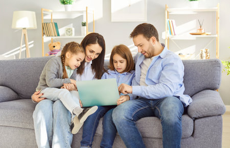 Happy mother, father, and two children spending leisure time at home on a couch, enjoying online entertainment using a laptop. Family bonding, relaxation, and digital fun in internet.の写真素材