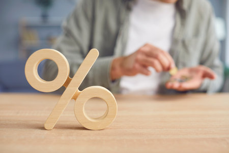 Close up cropped shot of wooden percentage symbol on desk. Young man counting money in his hands in the background. interest rate decline, investment reduce or sales discount concept. Selective focus.の写真素材