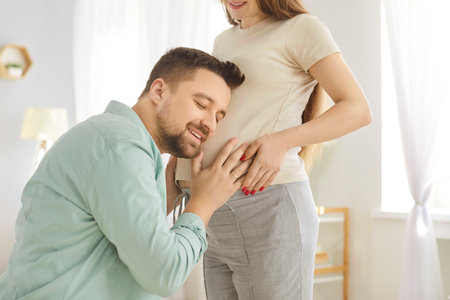 Happy loving husband listening to baby in pregnant wife belly in the living room at home. Young couple and future parents expecting a baby. Parenthood, maternity and pregnancy concept.の写真素材