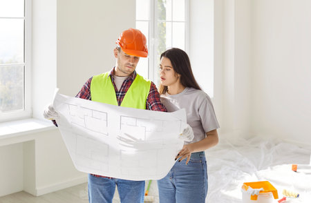 Foreman worker and female client watching flat plan to supervise and direct, worker looking at...