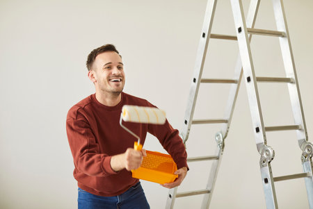 Smiling happy funny man homeowner having fun while painting the wall of new apartment holding paint roller in hand. Male person doing repair renovation and dancing preparing to move.の写真素材