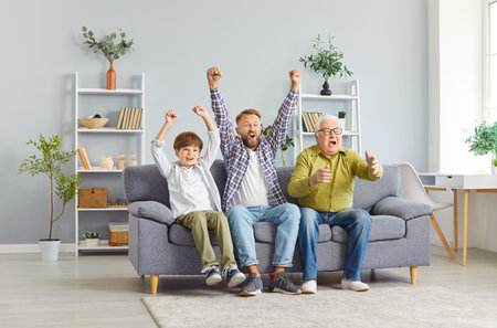 Excited senior grandfather, father and little child boy sitting on sofa at home watching football game on TV together. Family football fans spending time watching sport match. Family leisure concept.の写真素材