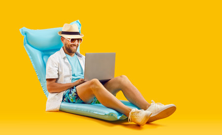 Happy man looking for sea tickets and booking hotel while sitting with laptop on inflatable mattress. Millennial man in beachwear on orange background is sitting on pool mattress and typing on laptop.の写真素材