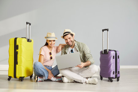 Happy couple travelers sitting on floor with suitcases and booking tickets for vacation trip via laptop on gray background. Young man and woman getting ready for holiday trip. Vacation concept.の写真素材