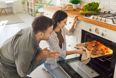 Happy smiling couple pulling baked muffins from oven in kitchen at home together. Young man and...