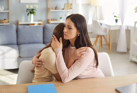 Loving mother hugging and comforting upset teenage daughter at table in living room. Worried parent embracing girl with care and concern, showing support and understanding during emotional moment.の写真素材