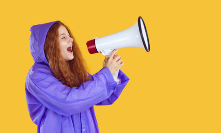 Funny woman in raincoat yelling in megaphone. Young long haired ginger girl in purple rain coat isolated on yellow background holding loudspeakerの写真素材