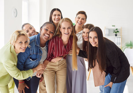 Group portrait of happy diverse women standing together in office and hugging each other. Happy female colleagues or friends laughing and embracing, showing unity and teamwork in positive atmosphere.の写真素材