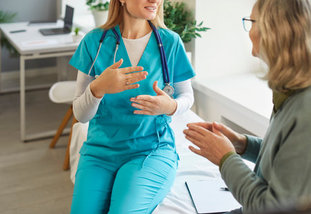 Cropped shot of young female doctor in medical uniform sitting on couch and explaining diagnosis to elderly patient. Senior woman gesturing while discussing treatment during consultation in clinic.の写真素材