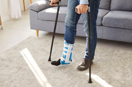 Midsection shot of a man with a broken leg in a cast walking with crutches at home during recovery....