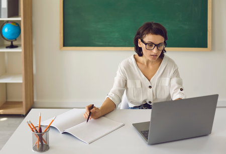 Serious school teacher focused on her online class. Young woman in glasses sitting at desk in classroom and using laptop computer to teach her students via video conferenceの写真素材