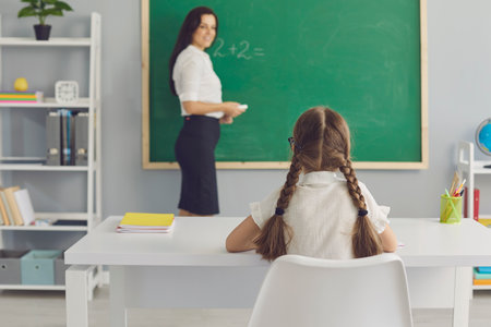 Pupil and teacher. Smart schoolgirl sitting at desk in modern classroom. Smiling tutor standing near blackboard waiting for her elementary student to do arithmetic sum. Primary education conceptの写真素材