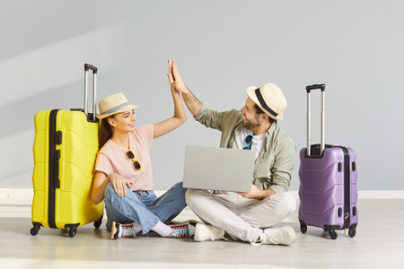 Happy husband and wife giving high five as reaching agreement where to travel, posing crossed legs on floor with laptop for making reservation online, packed suitcases for future tripの写真素材