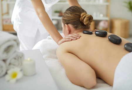 Young woman relaxing in spa salon getting massage therapy with hot stones from cosmetologist. Beautician therapist doing back treatment procedure. Wellness, body care and beauty day concept.の写真素材