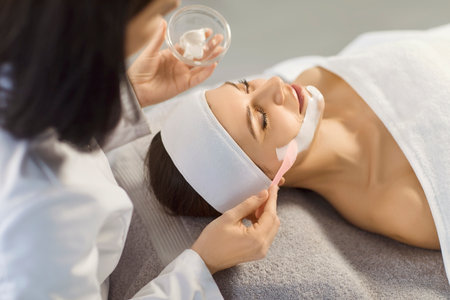 Cosmetologist applying moisturizing mask to a young relaxed smiling young woman in spa salon. Close up portrait of a girl client receiving beauty procedure from beautician in beauty clinicの写真素材