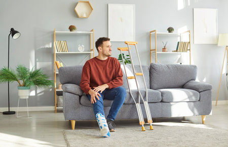 Young man with a leg injury resting at home during his sick leave. Upset man with a foot brace on his broken ankle sitting on the sofa with a pair of crutches besideの写真素材