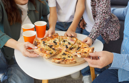 Close-up shot of a parents with two children in family setting eating fast food pizza for lunch together at home living room, enjoying leisure in a cozy environment with joy and connection.の写真素材