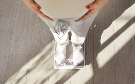 Cropped image of faceless pregnant woman standing on scales, controling her weight, touching belly, posing indoors. Weight gain during pregnancy. Unknown future mother preparing for child birthの写真素材