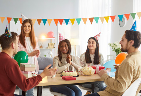 Company of friends meeting together to celebrate birthday, clapping hands and congratulating friend, wearing party hats, sitting at festive table with cakes. People and celebration conceptの写真素材
