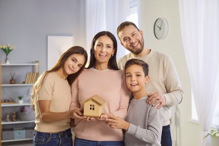 Portrait of happy joyful young family of father, mother and children holding wooden house in hands and looking cheerful at camera standing at home. Real estate and mortgage concept.の写真素材
