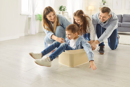 Happy parents playing with children in new apartment living room on floor, little children sitting in box laughing father pushing them for fun, family celebrating moving day, playful cheerful togetherの写真素材