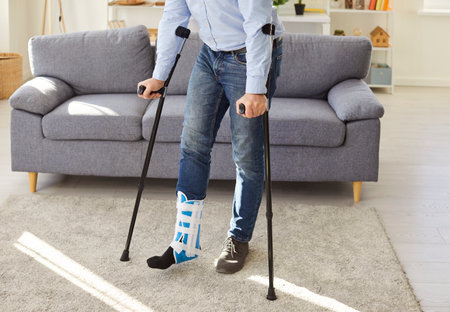 Cropped shot of man in orthopedic boot using crutches in bright living room. Injured person recovering from leg injury, balancing on crutches, showing determination and patience during rehabilitation.の写真素材