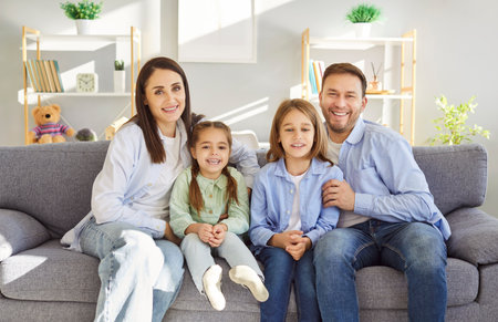 Happy loving family portrait, smiling bonding mother and father with two little children posing at home, looking at camera, family enjoying moment, children having fun together in modern living roomの写真素材