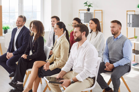 Group of smiling young business people sitting in a row on chairs and listening to colleague or their leadership at working place during business training or conference in meeting room.の写真素材
