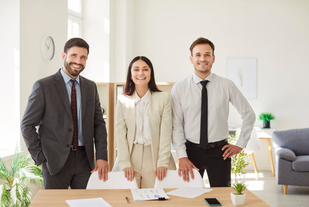 Group portrait of three professionals in formal wear standing by desk in bright office space, smiling at camera. Team of confident joyful colleagues two men and woman showing positive work atmosphere.の写真素材