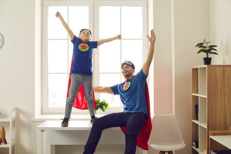 We can do it. Happy young dad and active kid dressed as superheroes enjoying time together. Funny father and child in super hero cloaks playing games at home. Fun childhood, parents support conceptの写真素材