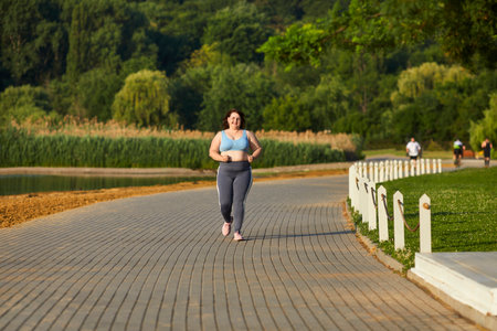 Happy smiling woman running in the summer park in nature. Plus size girl wearing sportswear jogging outdoors. Body positive, sport and fitness lifestyle concept.の写真素材
