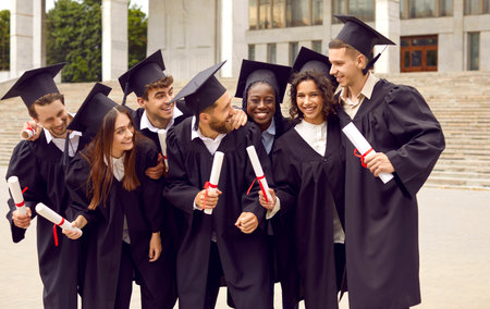 Group of happy successful multiracial students celebrating university graduation day together. Students in academic gowns laughing standing near university stairs. Education and graduation concept.の写真素材