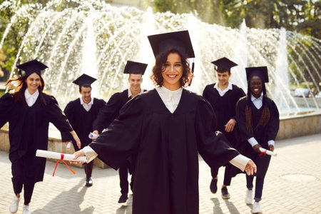 Young smiling joyful girl student in university graduate gown and diploma in hands. Happy graduated woman standing with her classmates outdoor. Graduation celebration and education concept.の写真素材