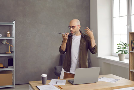 Serious professional business man on speakerphone. Bald middle aged entrepreneur standing by working desk table with notebook PC in home office, holding phone, recording thought, idea or voice messageの写真素材