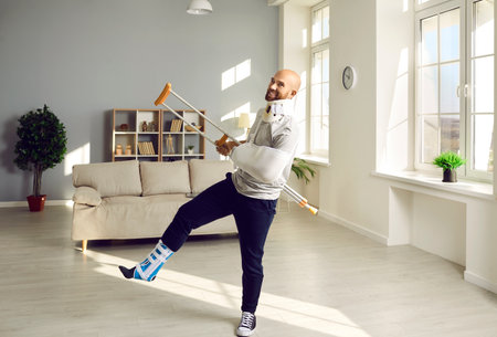 Man has fun during rehabilitation at home recovering from numerous physical injuries. Smiling young man with leg, arm and neck injury stands in living room on one leg with crutch raised.の写真素材