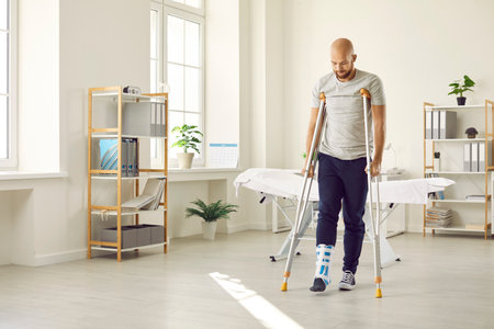 Orthopaedic recovery. Man wearing support adjustable fracture fixator on injured leg walks on crutches in hospital office. Concept of rehabilitation of people after serious physical accident injury.の写真素材